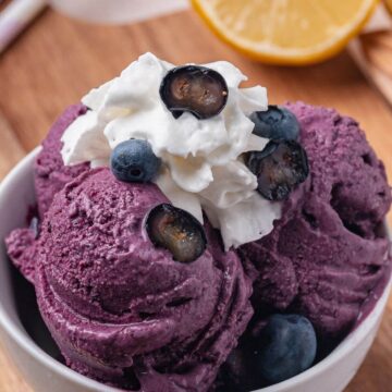 A bowl of blueberry ice cream topped with whipped cream and fresh blueberries, with a sliced lemon in the background.