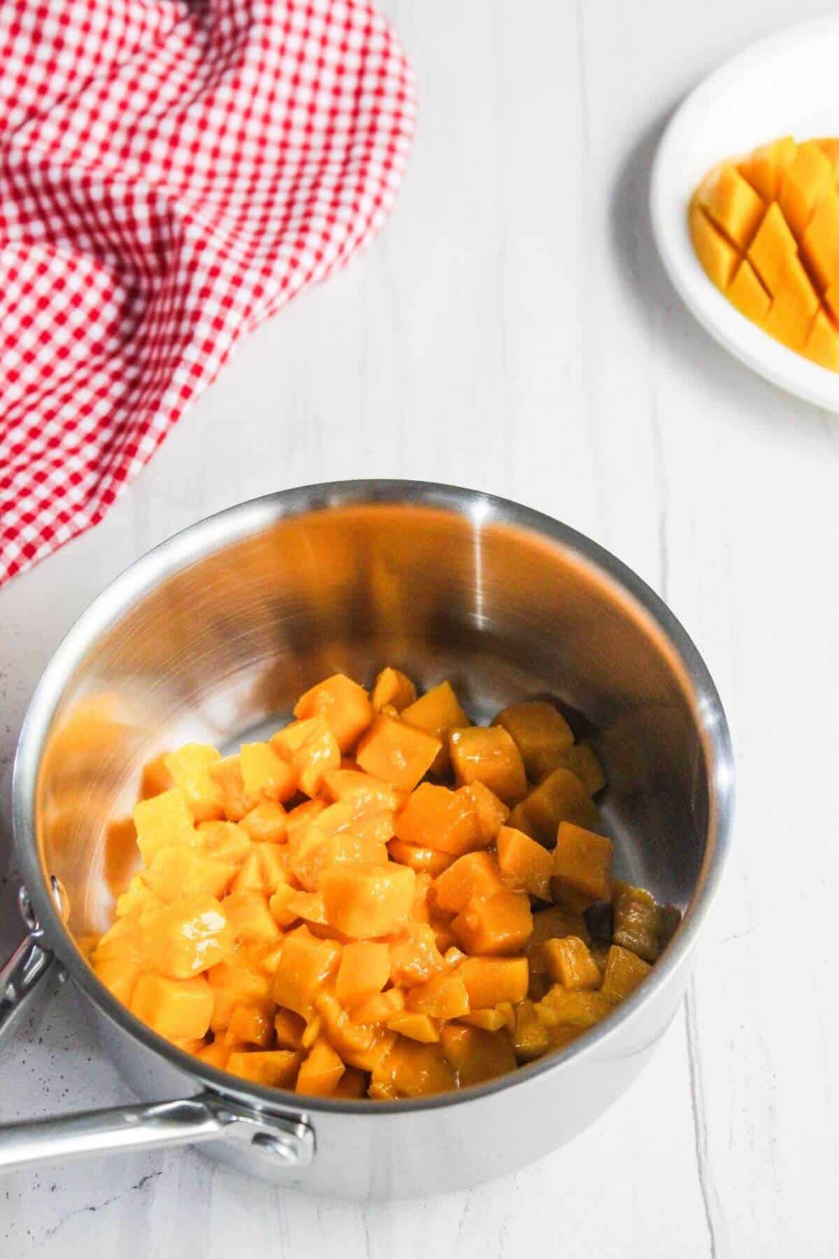 A stainless steel pot filled with diced mango sits on a white surface, with a plate of more diced mango and a red and white checkered cloth nearby.