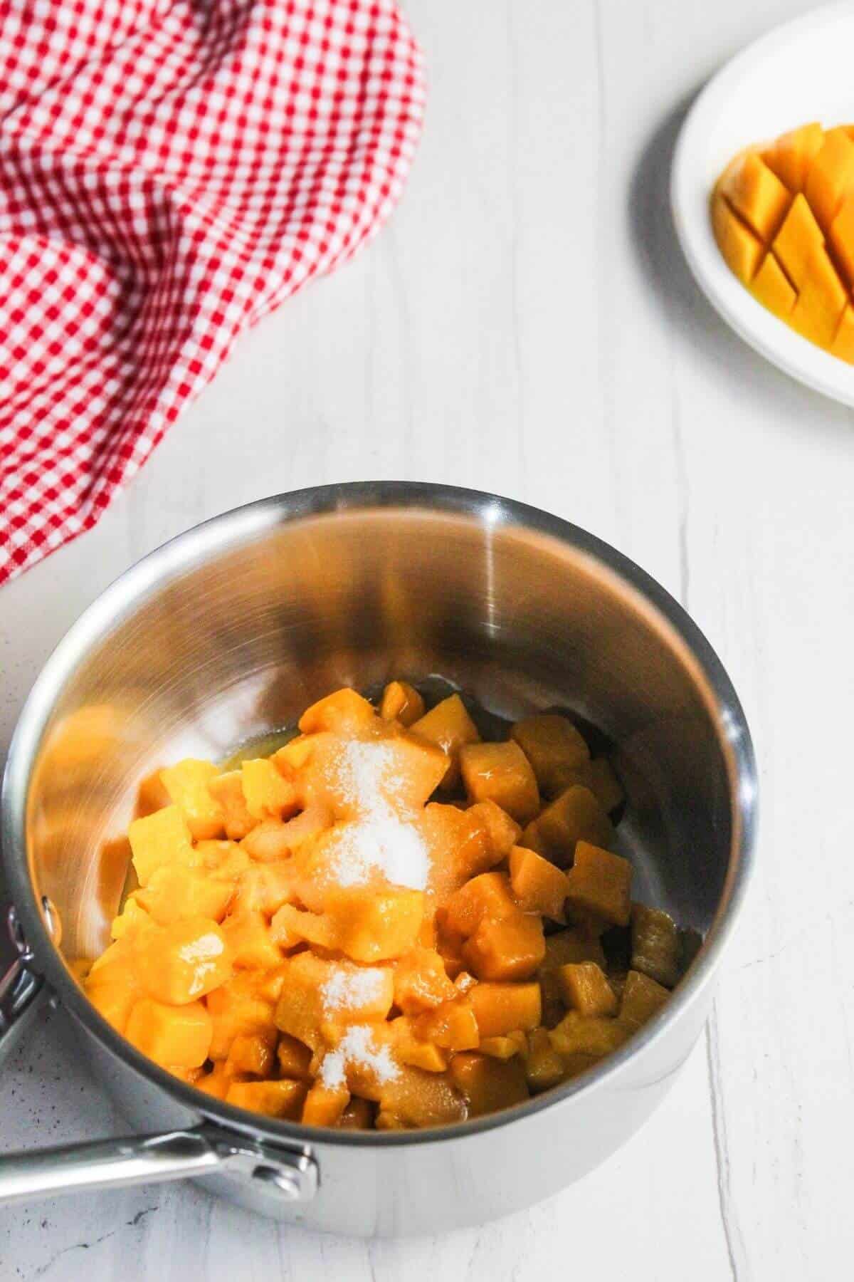 A stainless steel pot with diced mango and sugar inside sits on a white surface, with a red checkered cloth and a plate of diced mango in the background.