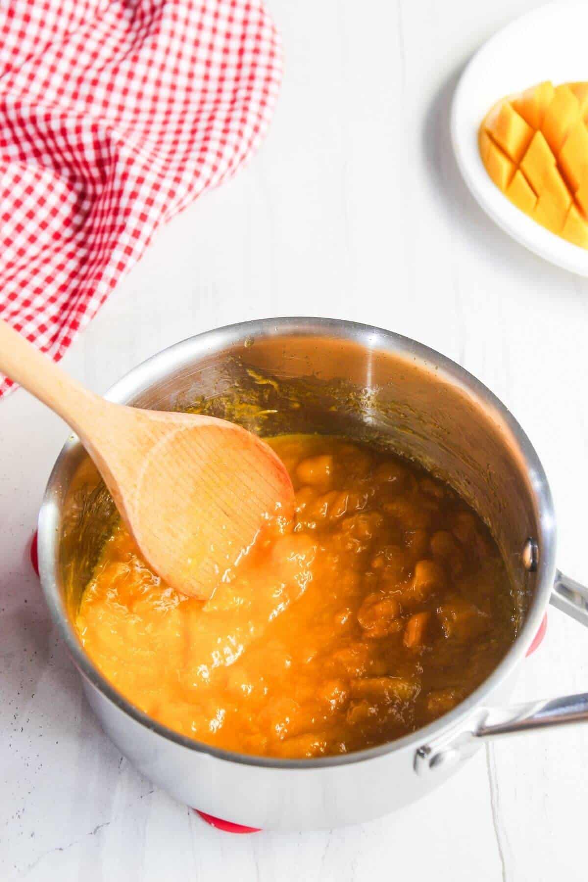 A wooden spoon stirring mango puree in a saucepan on a white countertop, with a sliced mango and red checkered cloth nearby.