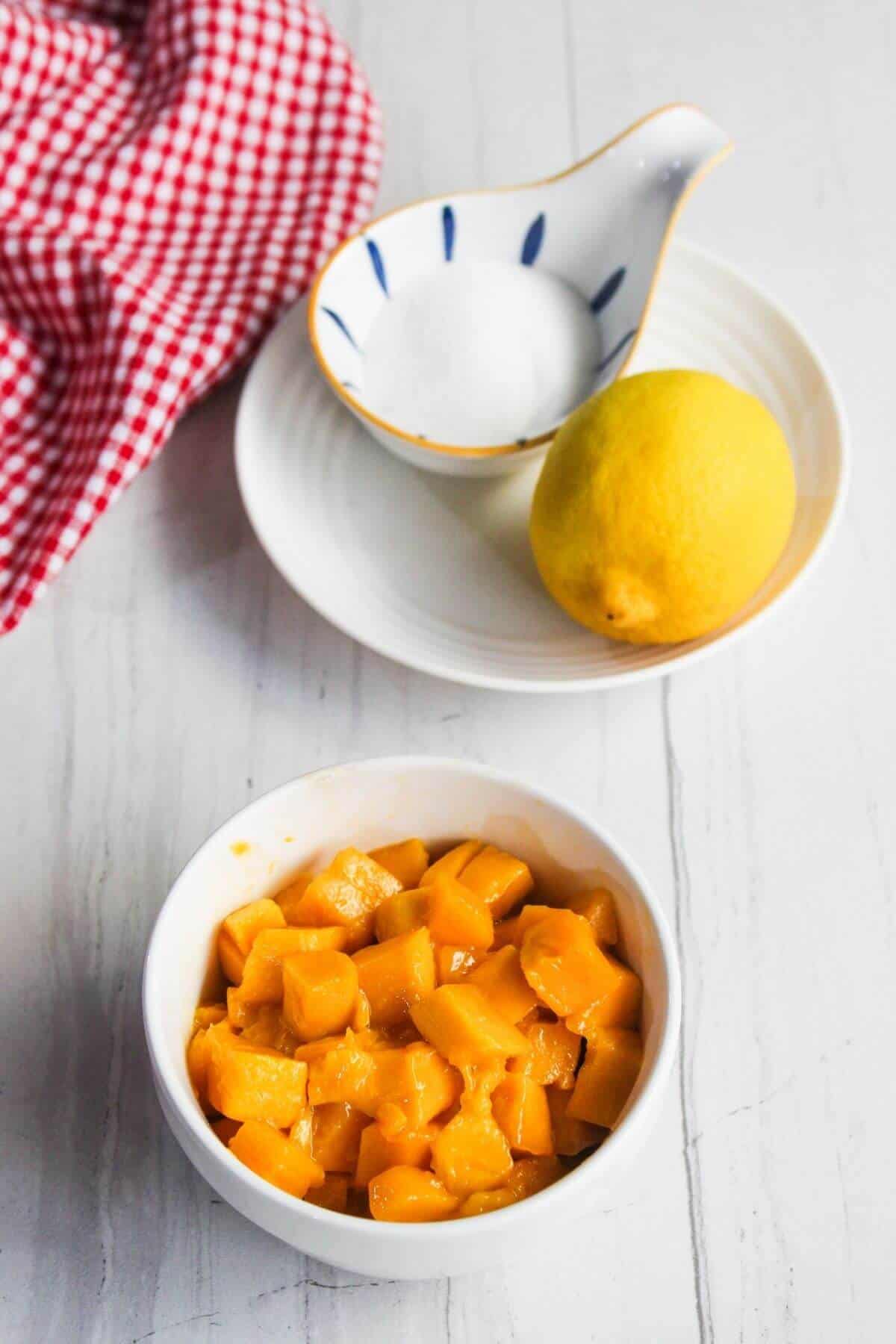 A bowl of diced mango, a lemon, and a small dish of sugar are arranged on a white surface next to a red and white checkered cloth.