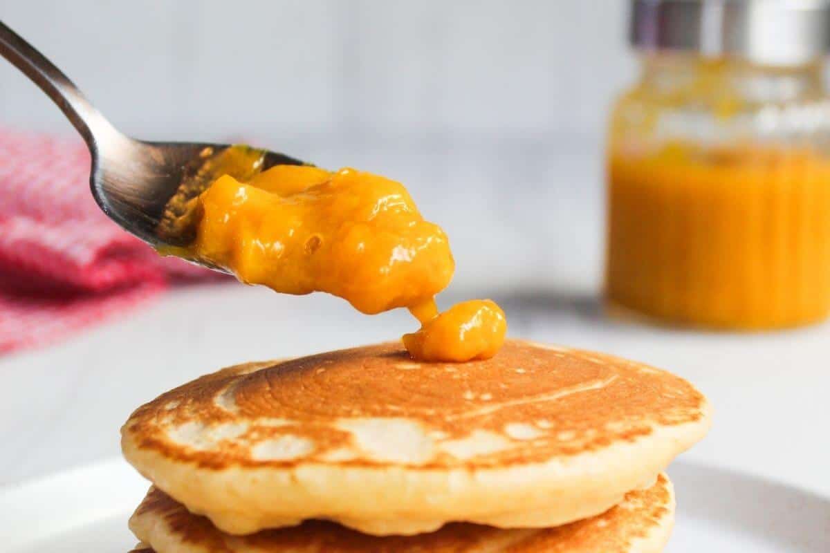 A spoonful of orange sauce is held above a stack of pancakes, with a jar of the same sauce blurred in the background.