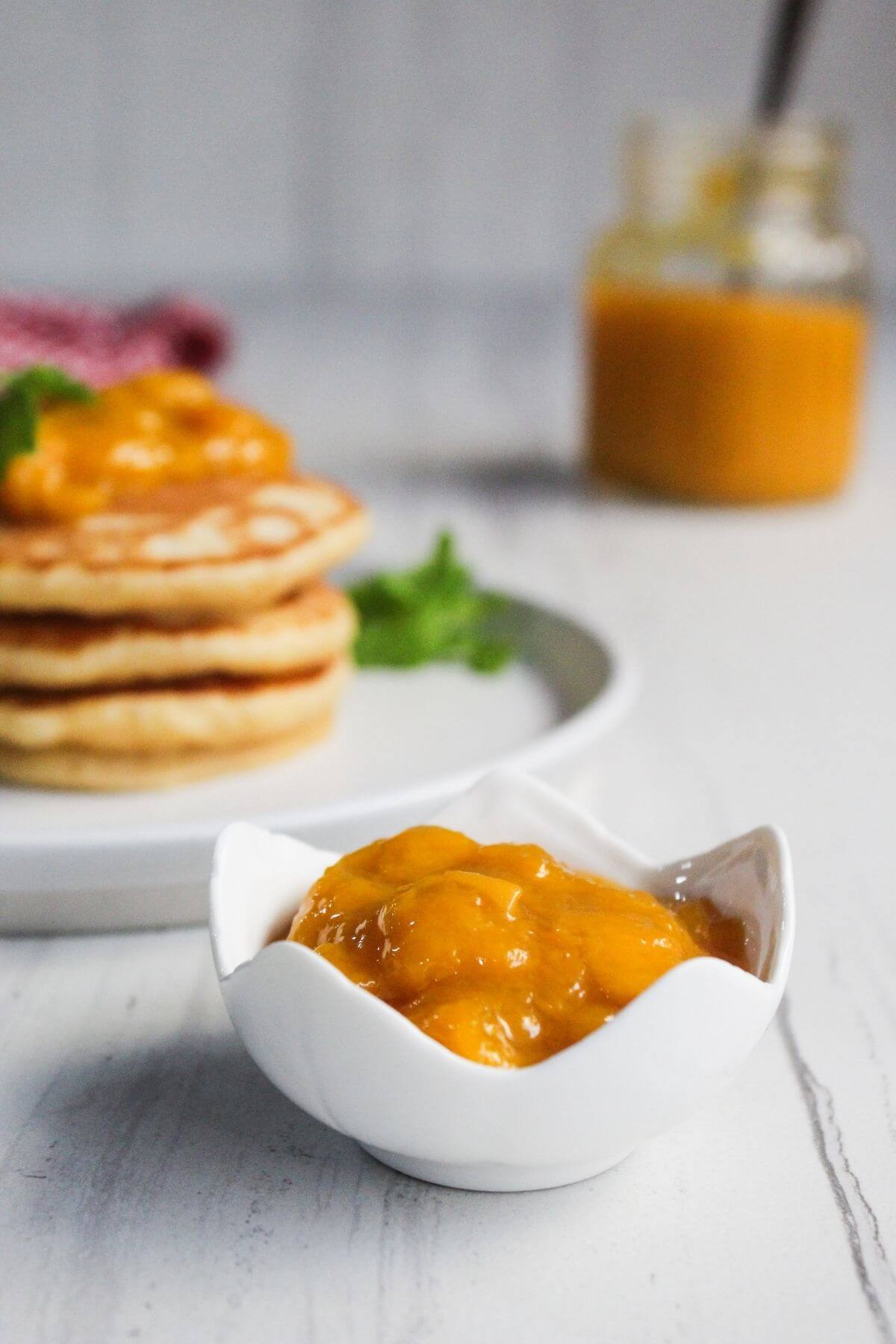 A small white bowl filled with mango chutney sits in the foreground, with a plate of pancakes and a jar of chutney blurred in the background.