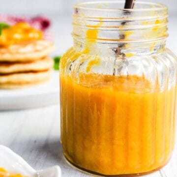 A glass jar filled with orange-colored fruit sauce sits on a table with a spoon inside; blurred pancakes are in the background.