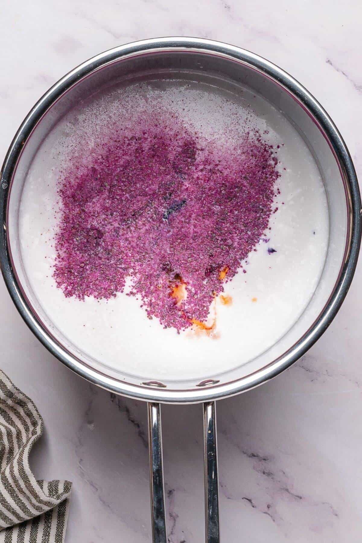 A saucepan contains white liquid with purple powder and a small amount of orange liquid on the surface, placed on a marble countertop.
