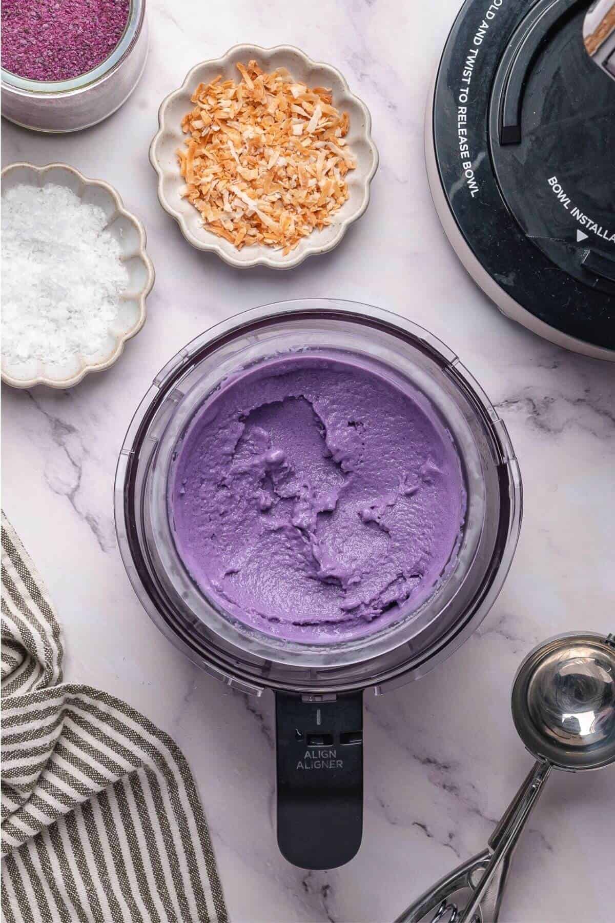 A food processor with smooth purple mixture inside, surrounded by bowls of shredded coconut, salt, and purple powder on a marble countertop.