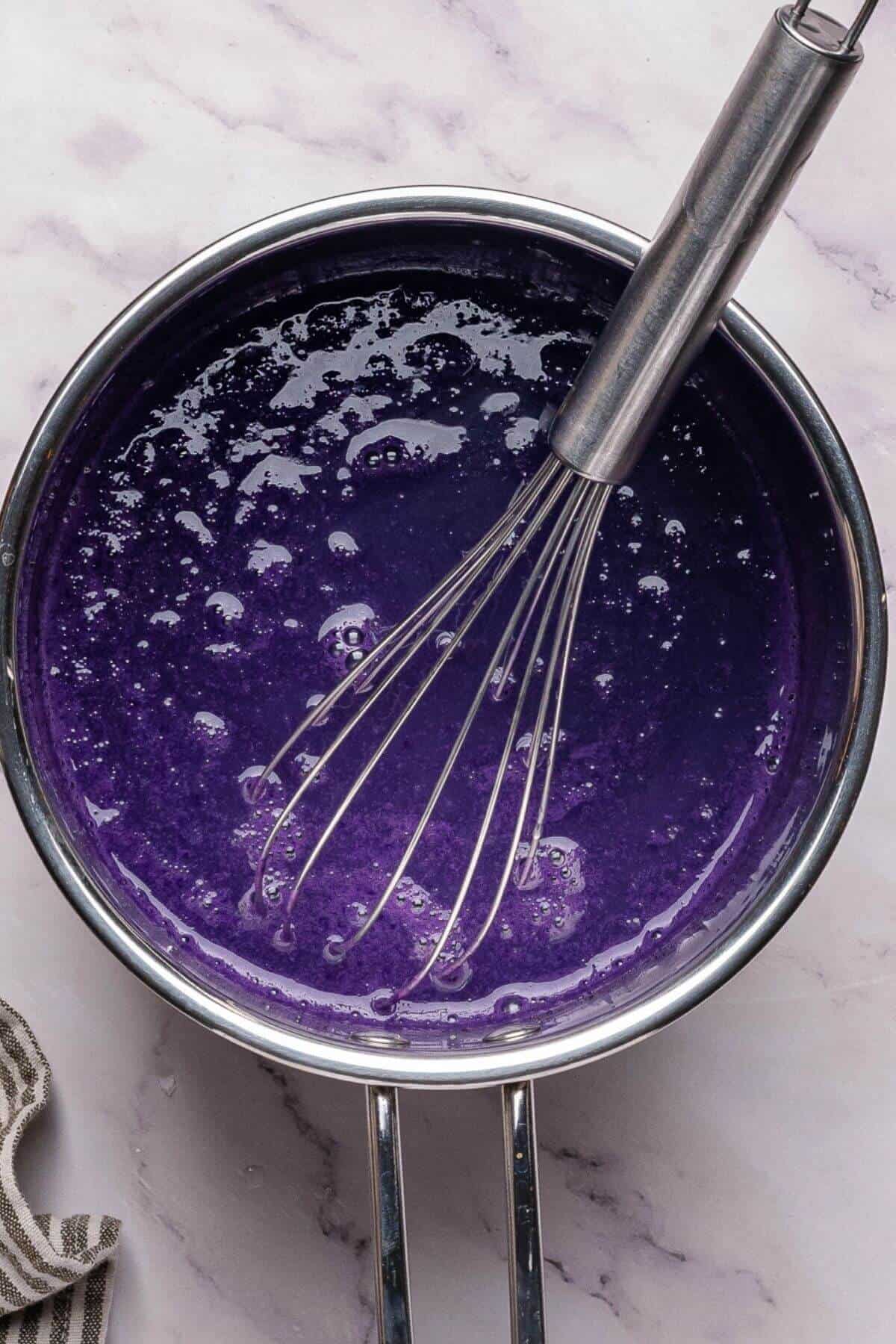 A saucepan filled with a bright purple liquid mixture and a metal whisk resting inside, on a marble surface.