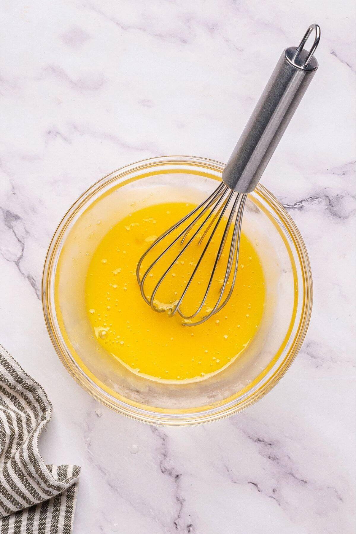 A glass bowl filled with a yellow liquid being whisked with a metal whisk, placed on a white marble surface next to a striped cloth.