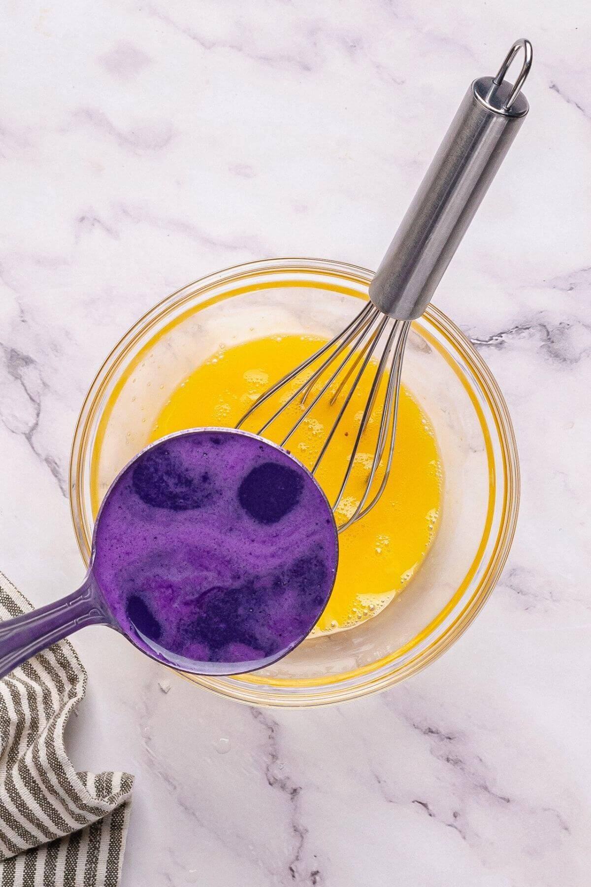 A glass bowl with yellow liquid and a whisk beside a striped cloth. A purple ladle with dark spots is placed above the bowl against a marble surface.