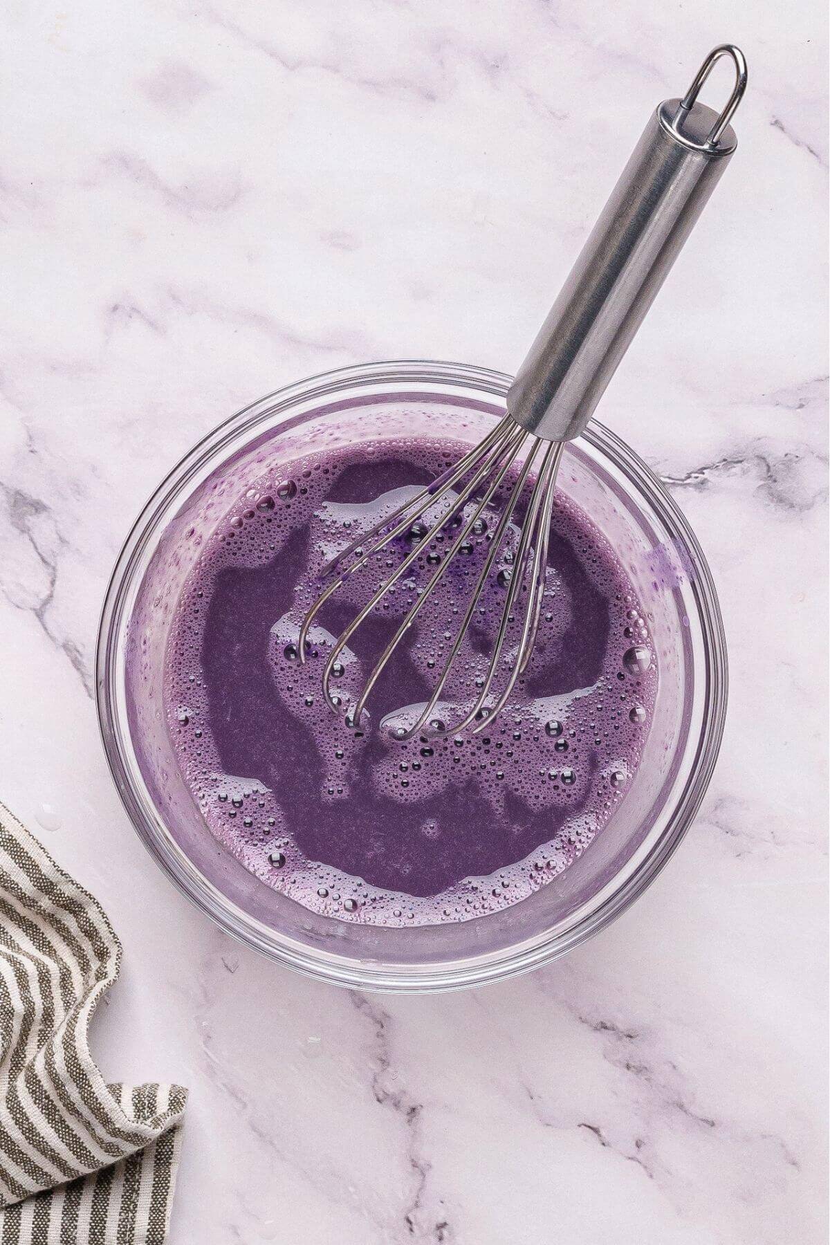 A glass bowl filled with purple liquid being whisked with a metal whisk, placed on a marble countertop next to a striped cloth.