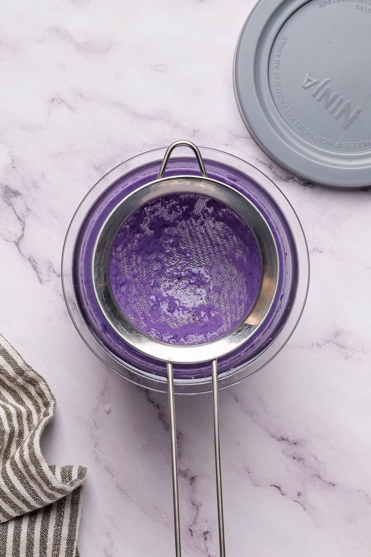 A metal strainer sits over a glass bowl containing a smooth purple mixture, on a marble surface next to a striped cloth and a gray lid.