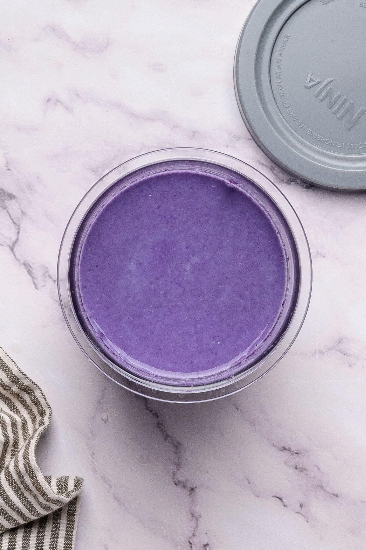 A clear container filled with smooth, purple ube halaya on a light marble surface, lid and striped cloth nearby.