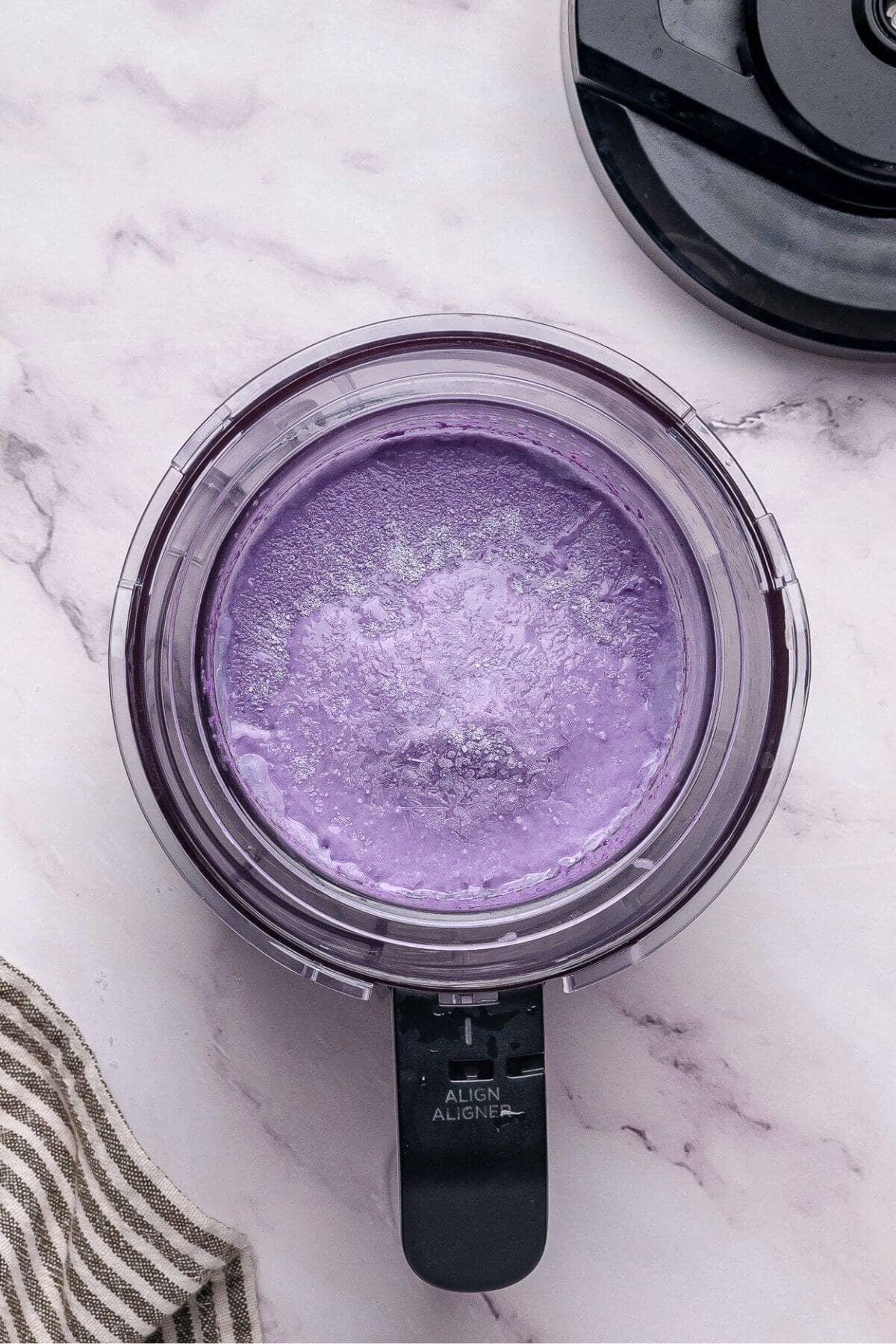 A food processor bowl filled with smooth, light purple cream sits on a marble surface next to a striped towel.