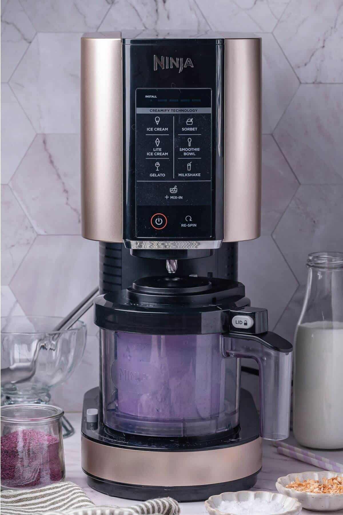 A Ninja ice cream maker with a transparent container of purple ice cream sits on a counter beside a glass of milk, a bowl of powder, and a metal scoop.