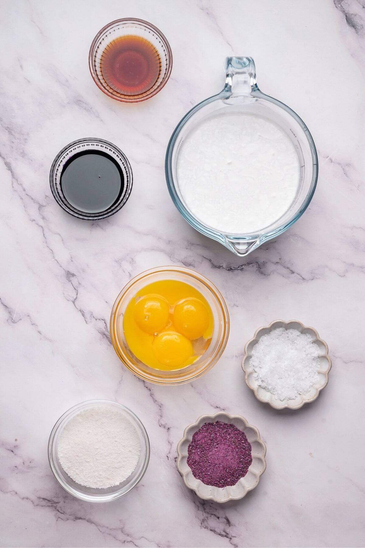 Assorted baking ingredients on a marble surface, including eggs, milk, vanilla extract, sugar, salt, a dark liquid, and a bowl of purple powder.