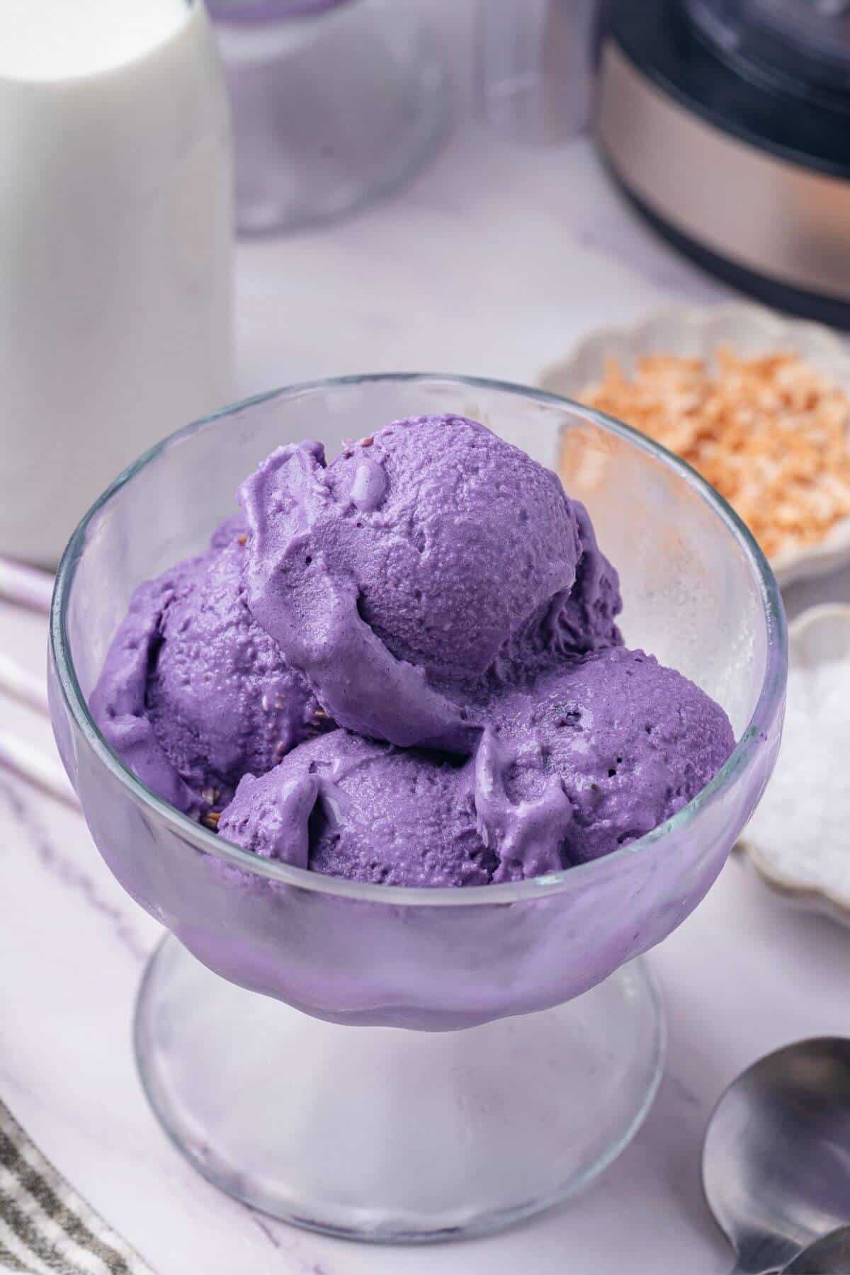 A glass bowl filled with three scoops of purple ube ice cream sits on a light surface, with a glass of milk and small dishes in the background.
