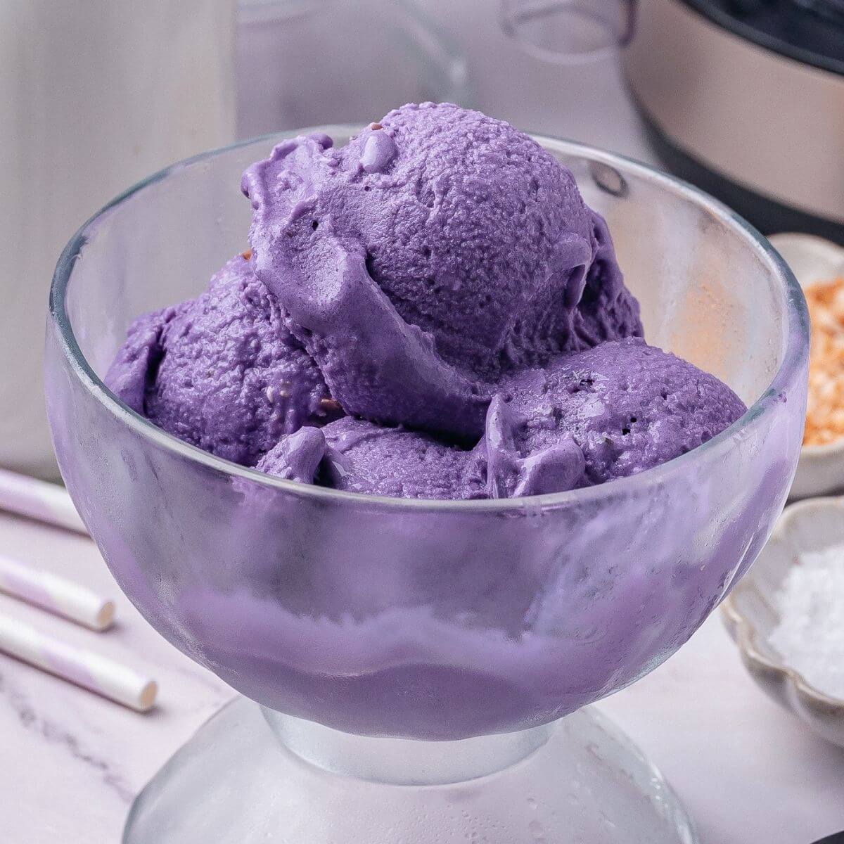 A glass bowl filled with several scoops of purple ice cream, set on a light-colored surface with blurred kitchen items in the background.