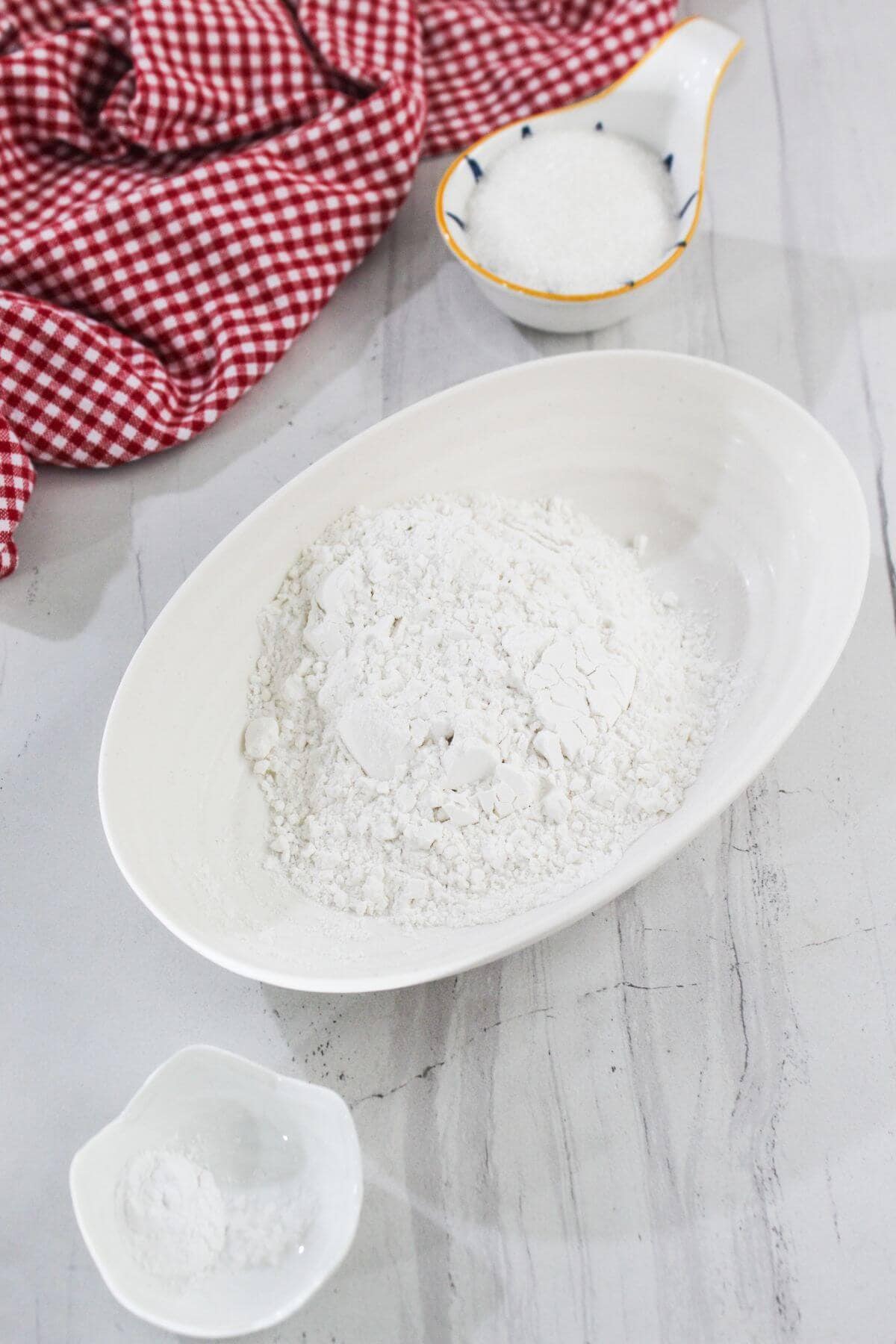 A white oval dish with flour, a small bowl of baking powder, and a small jug of granulated sugar on a light gray countertop with a red checkered cloth.