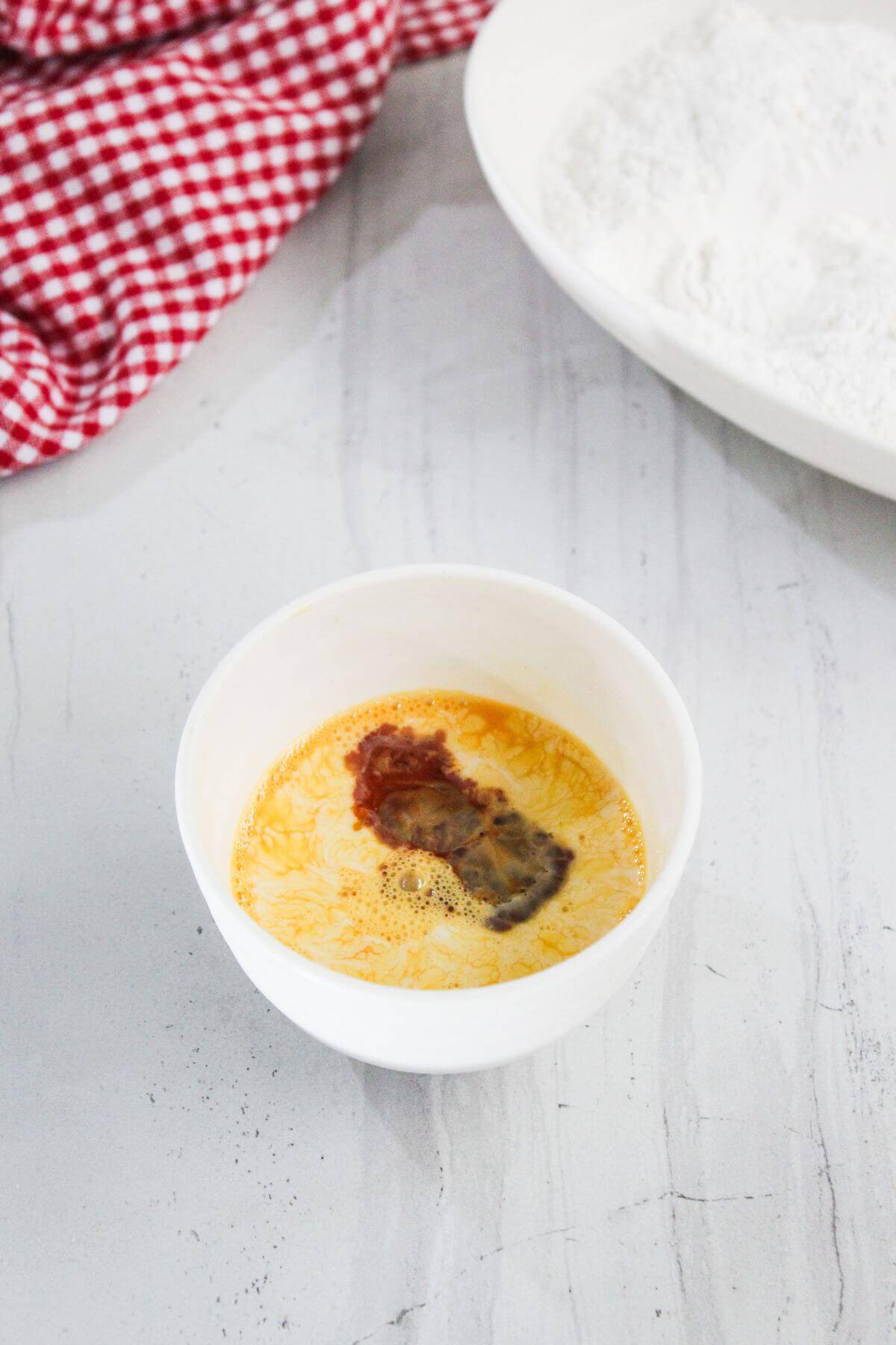 A white bowl with beaten eggs and a splash of soy sauce sits on a light countertop near a plate of flour and a red checkered cloth.