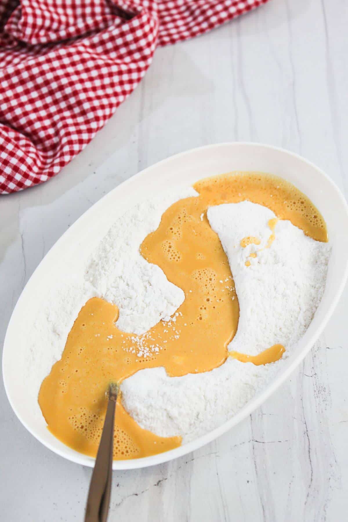 An oval dish with flour and beaten eggs being mixed, set on a white countertop with a red and white checkered cloth nearby.