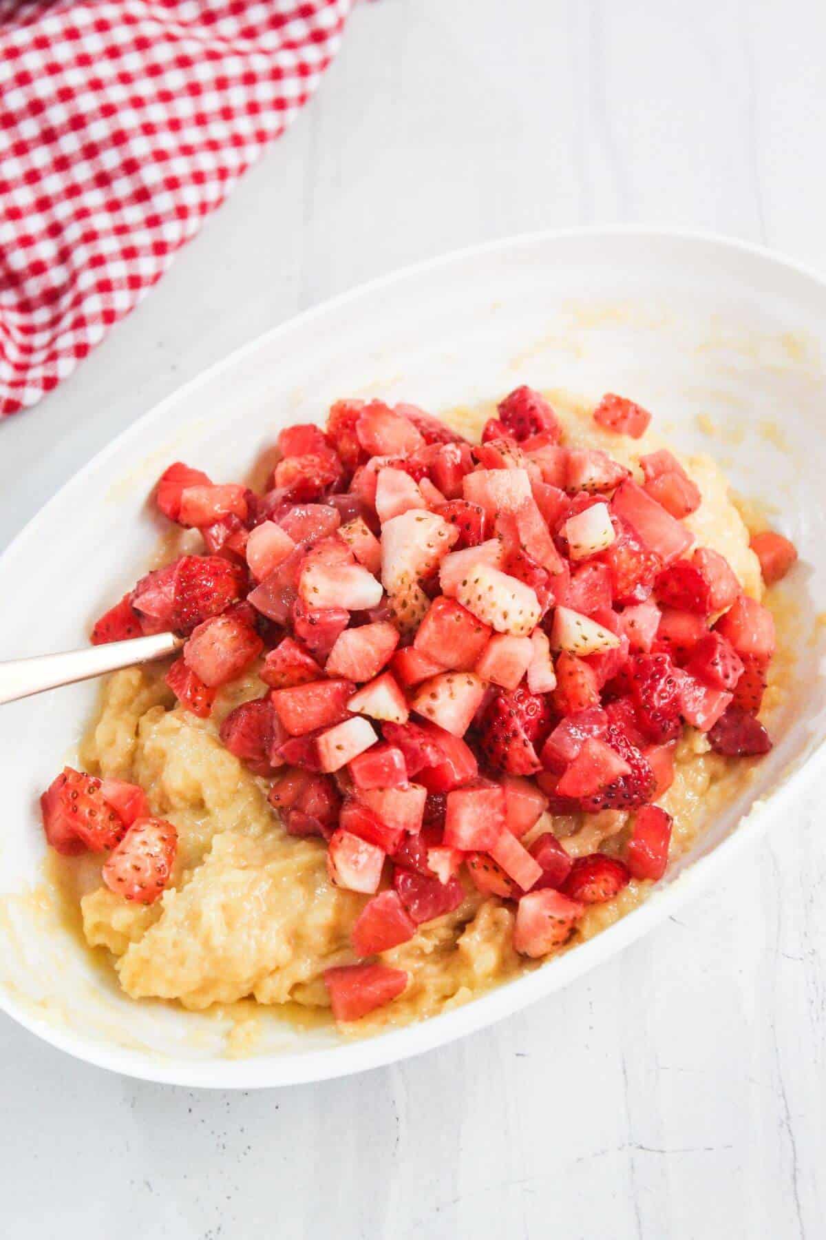 A white oval dish containing a mixture of mashed bananas and chopped fresh strawberries, with a spoon partially visible. A red and white checkered cloth is in the background.