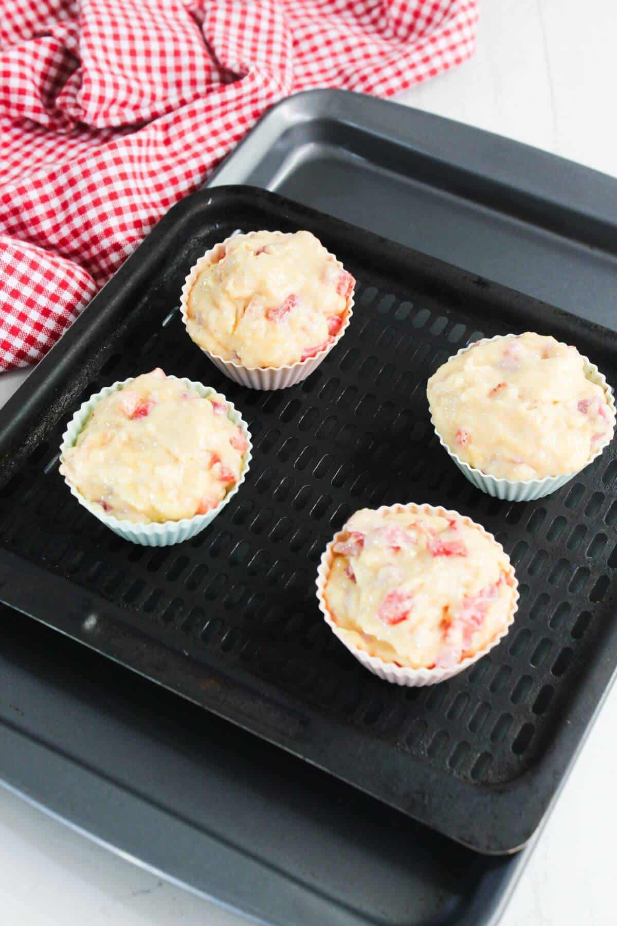 Four unbaked muffin batter portions in silicone cups on a black baking tray, with a red and white checked cloth in the background.