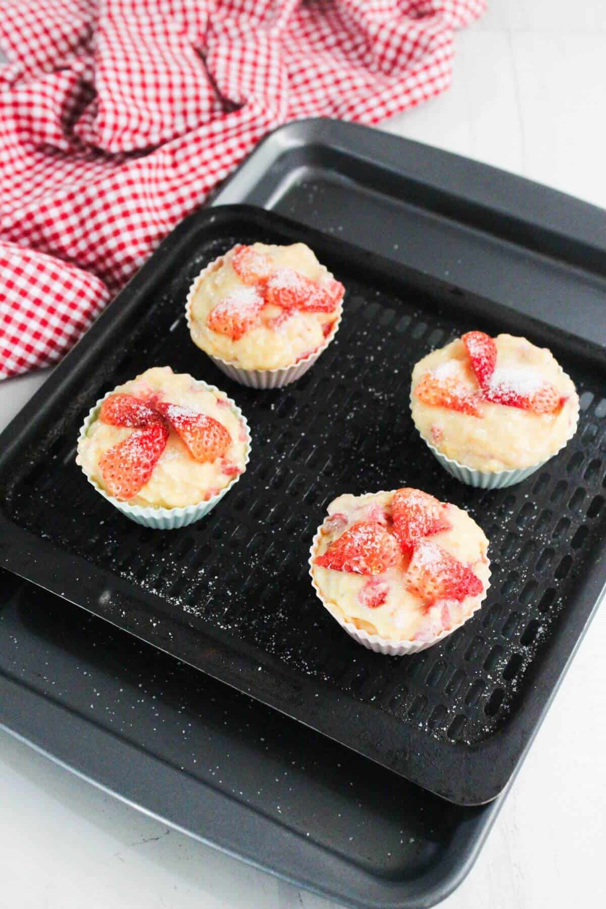 Four strawberry-topped cupcakes in paper liners are arranged on a black baking tray, with a red and white checkered cloth in the background.