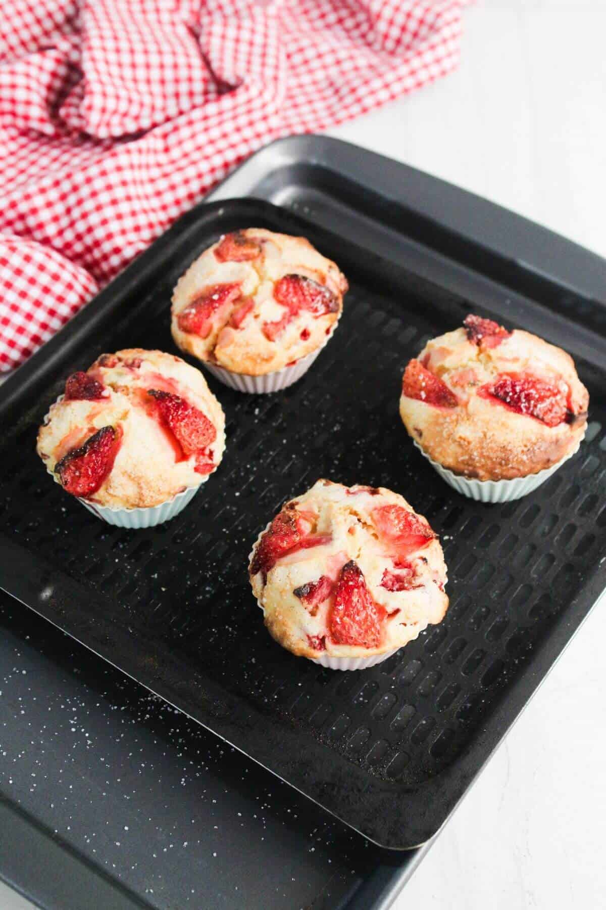 Four strawberry muffins in paper cups on a black baking tray, with a red and white checkered cloth in the background.