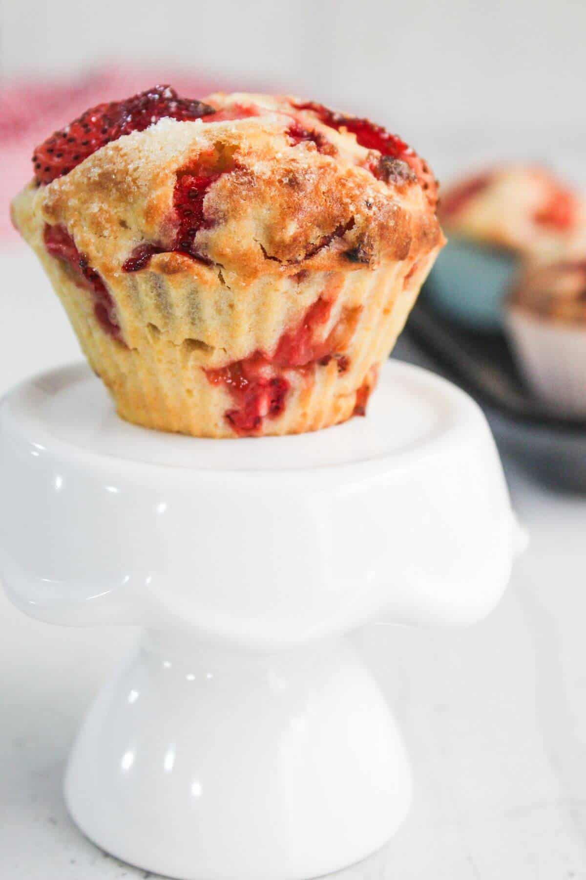A strawberry muffin in a silicone baking cup sits on a white pedestal, with another muffin blurred in the background.