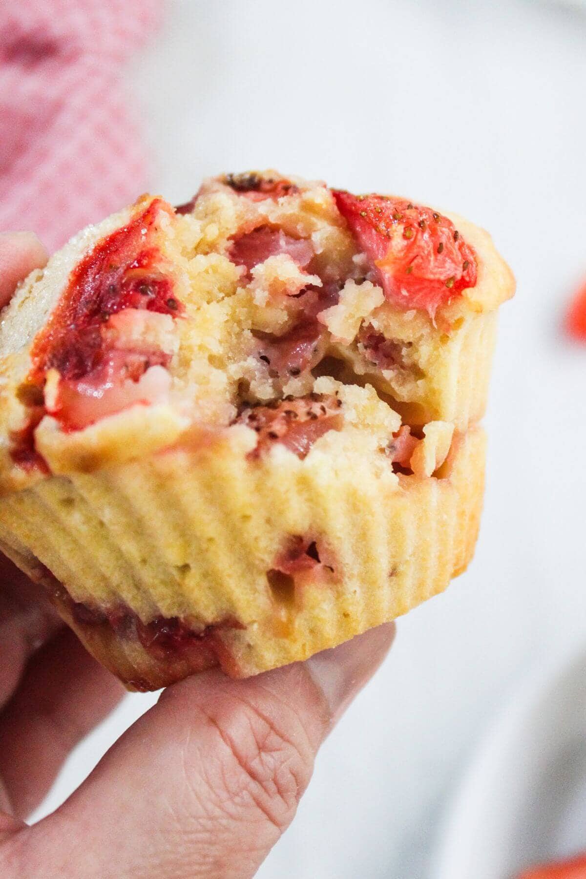 A hand holds a partially eaten strawberry muffin in a yellow paper liner, showing the moist interior and pieces of strawberry.