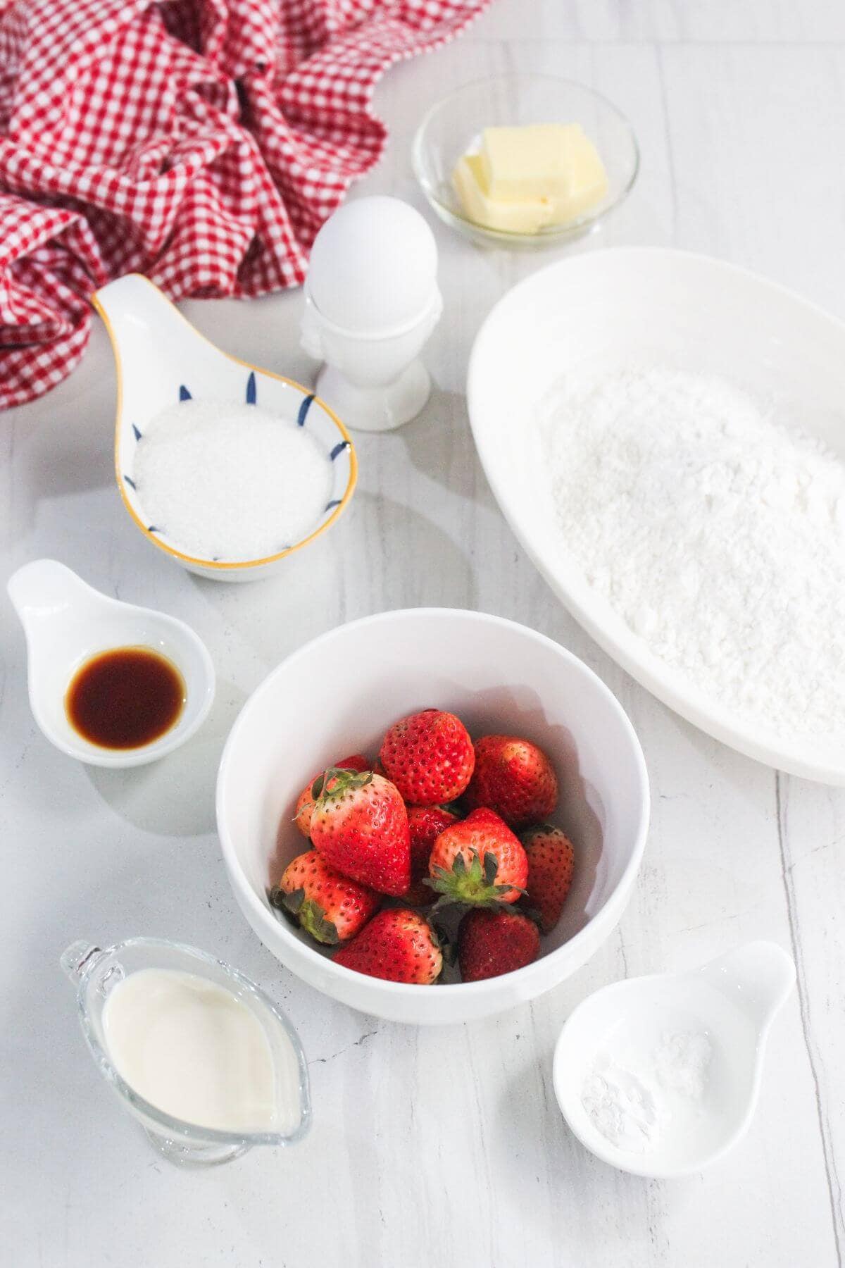 A bowl of strawberries, flour, butter, sugar, an egg, vanilla extract, milk, and baking powder on a white surface with a red checkered cloth.