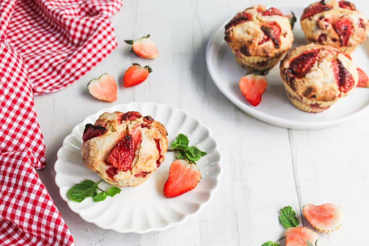 A strawberry muffin on a white plate, garnished with strawberry slices and mint, with more muffins and a red checkered cloth in the background.