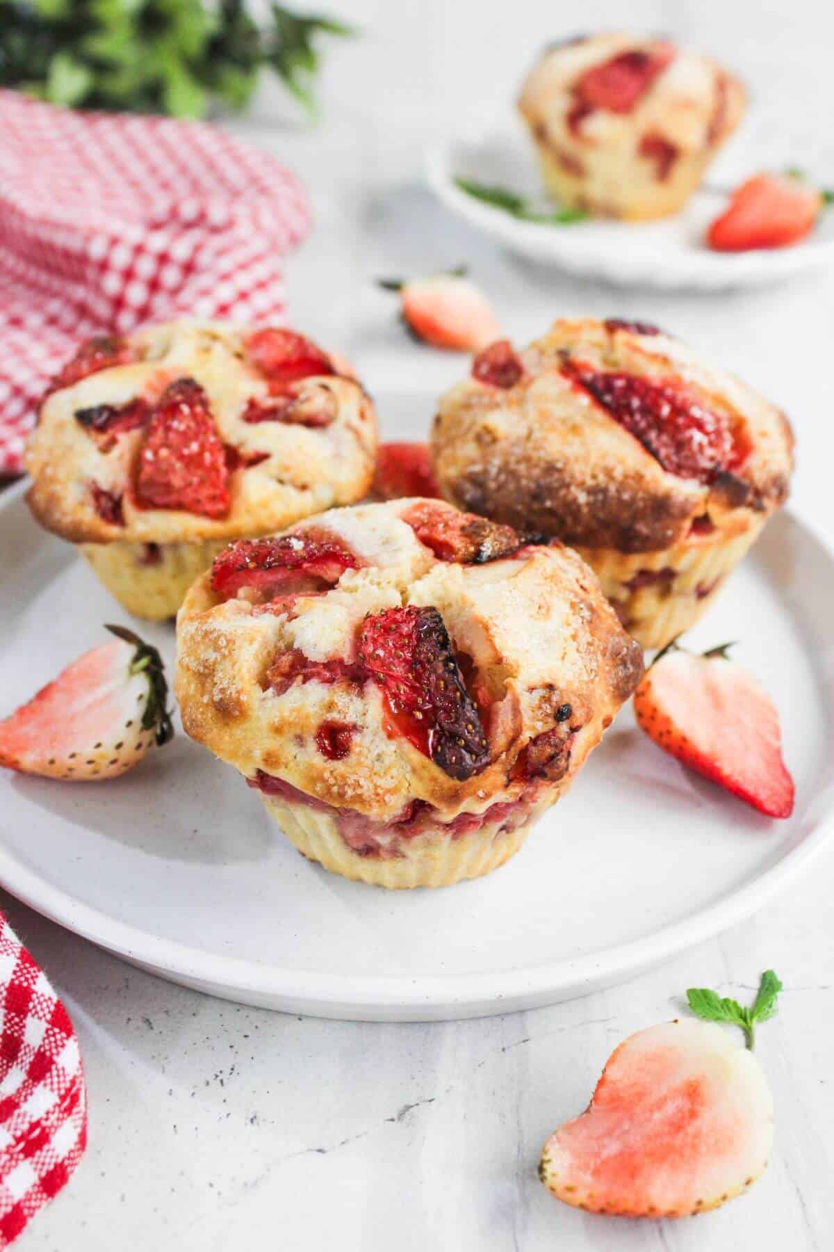 Three strawberry muffins on a white plate, surrounded by fresh strawberry halves, with a red checked cloth and another muffin in the background.