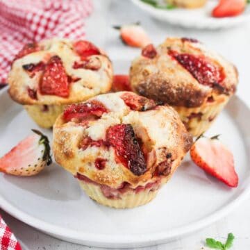 Three strawberry muffins with visible strawberry pieces are arranged on a white plate, surrounded by fresh strawberry halves.