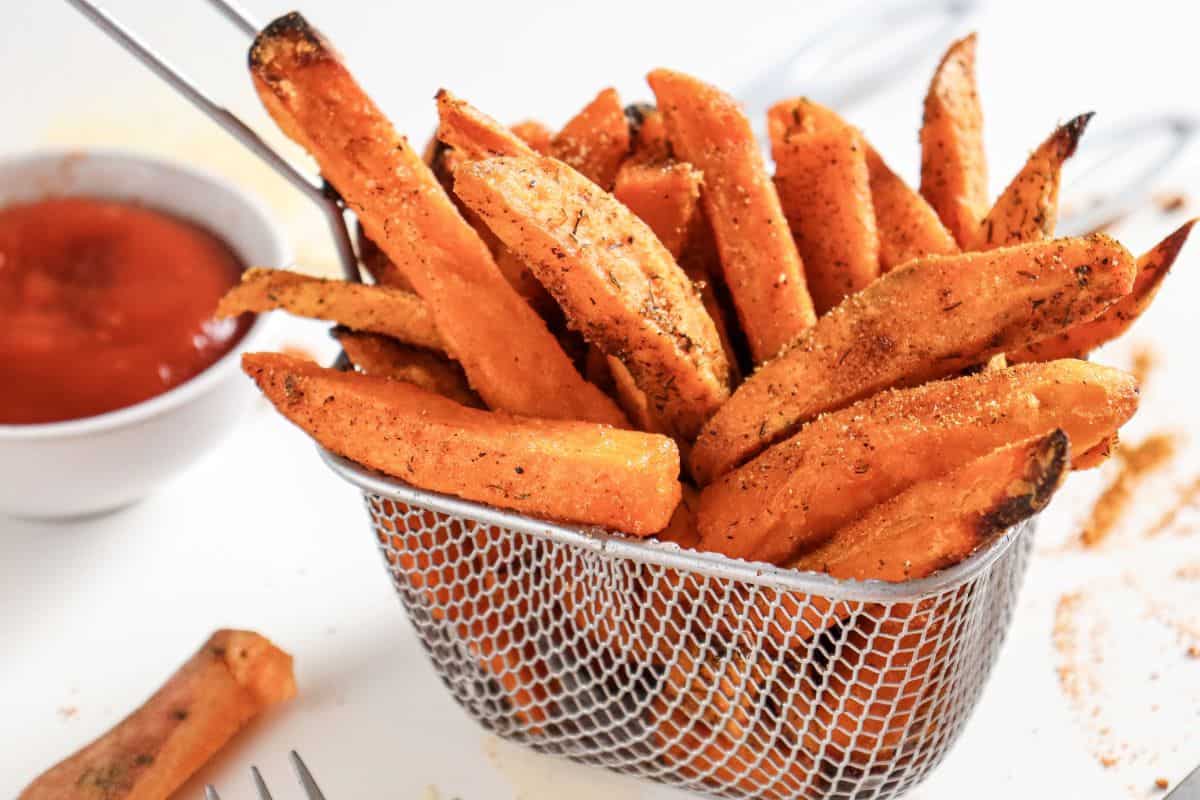 A metal basket filled with seasoned sweet potato fries sits on a white surface next to a bowl of red dipping sauce.
