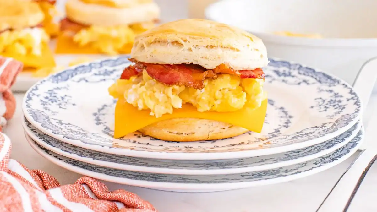 A breakfast sandwich with scrambled eggs, cheddar cheese, and bacon on a biscuit sits on a patterned plate.