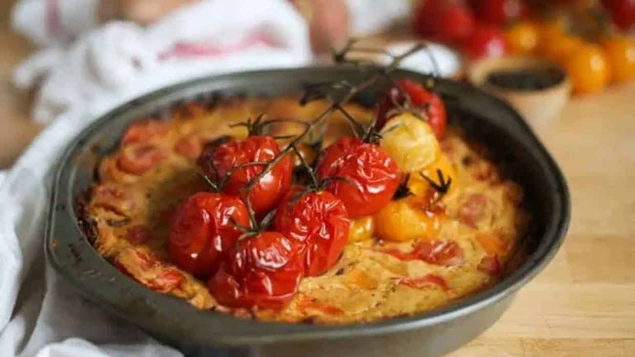 A baked frittata topped with roasted vine tomatoes sits in a round metal pan on a wooden surface.