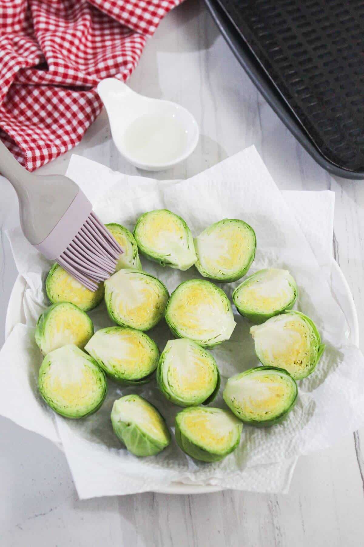 Halved Brussels sprouts on a paper towel-lined plate are being brushed with oil, with a red checkered cloth and a black tray nearby.