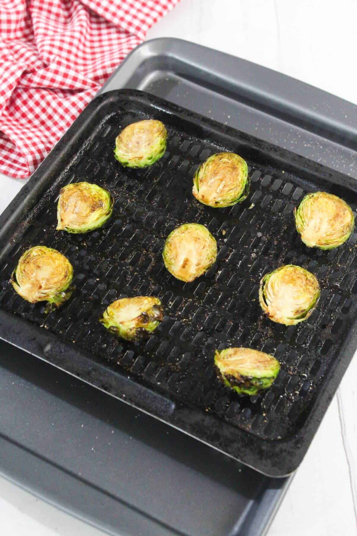 Halved Brussels sprouts arranged on a black grill tray, roasted and lightly browned, with a red and white checkered cloth in the background.