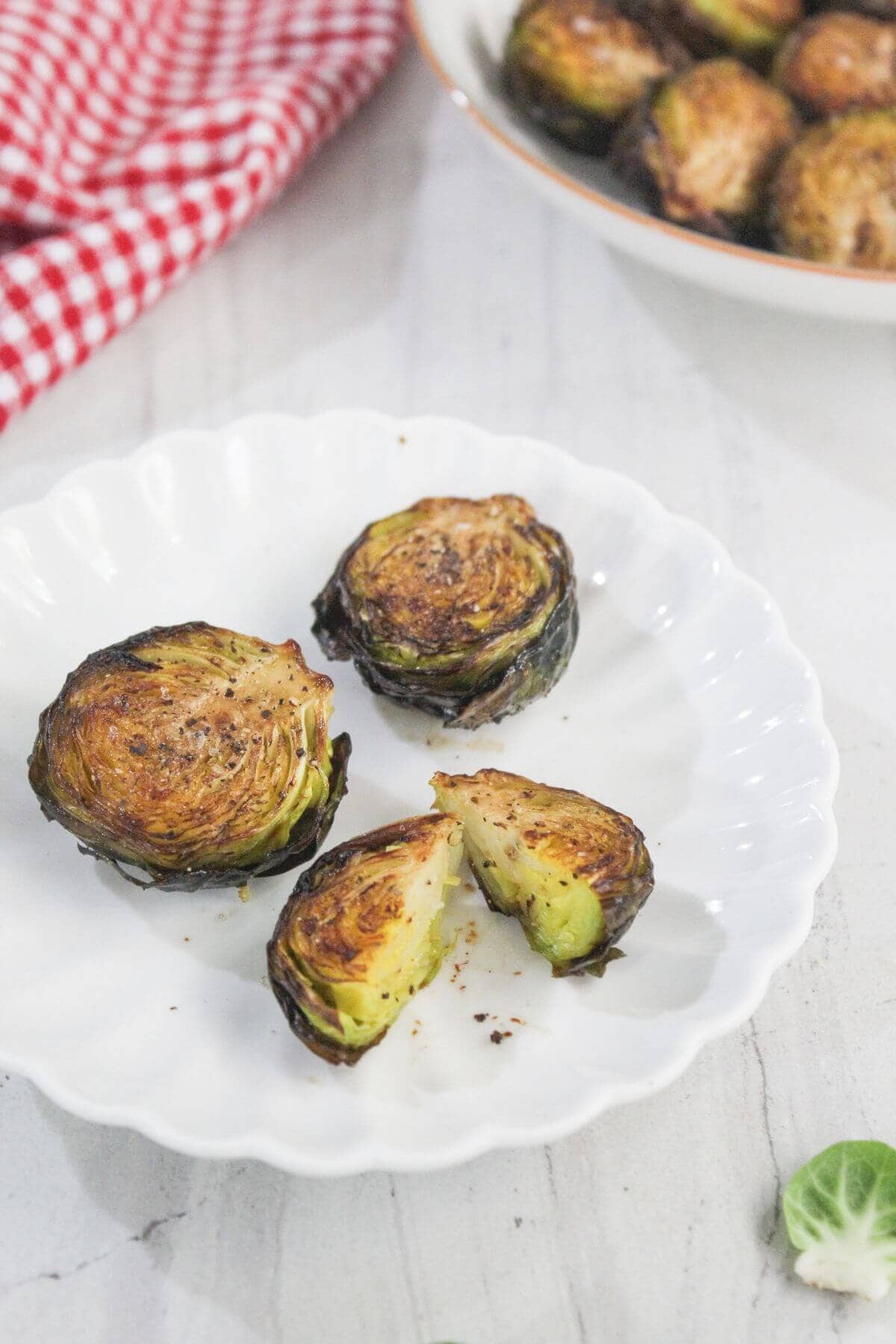 Four roasted Brussels sprouts, some halved, are arranged on a white scalloped plate. A red checkered cloth and a bowl of more sprouts are in the background.