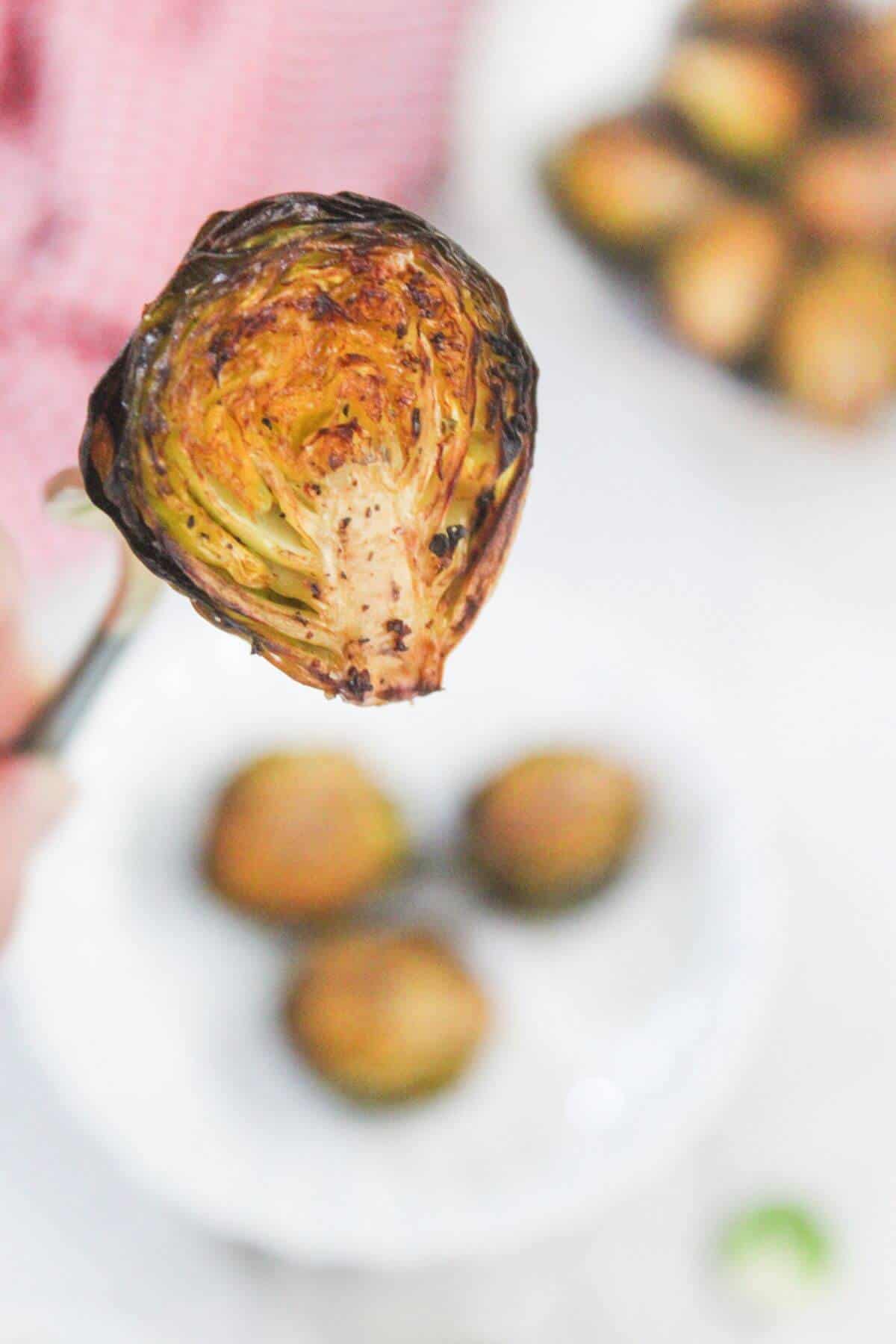 A roasted Brussels sprout held up by tongs, with more Brussels sprouts blurred in the background on a white plate.