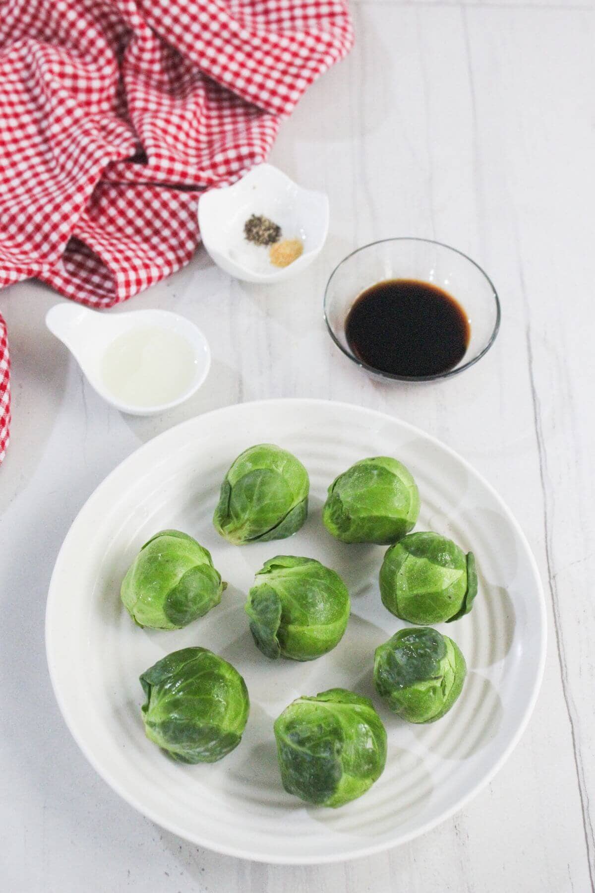 A plate with eight raw Brussels sprouts, a glass bowl of dark sauce, a small bowl of mixed spices, a small white pitcher of liquid, and a red checkered cloth on a white surface.