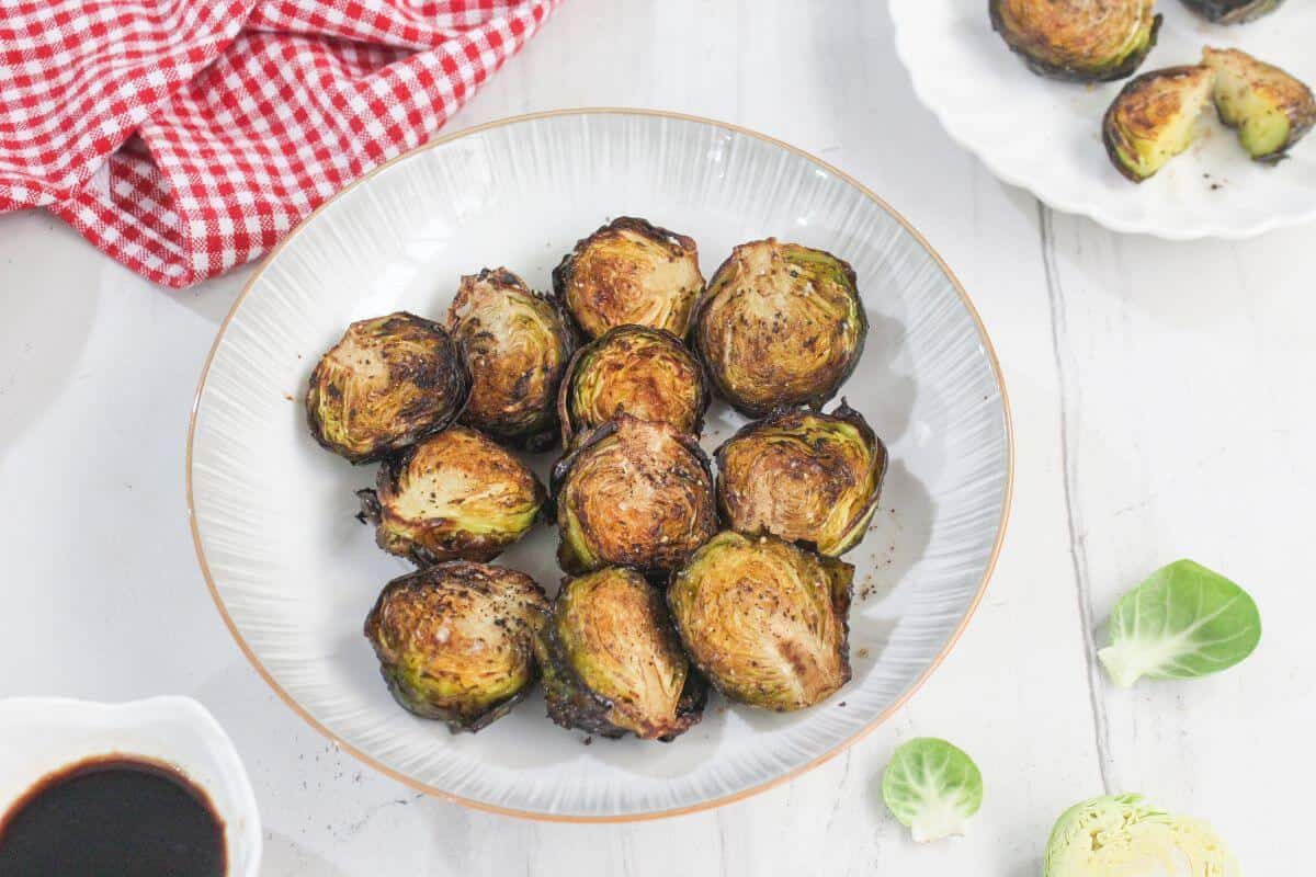 A white bowl filled with roasted Brussels sprouts sits on a white surface, with a red checkered cloth and a small bowl of dark sauce nearby.
