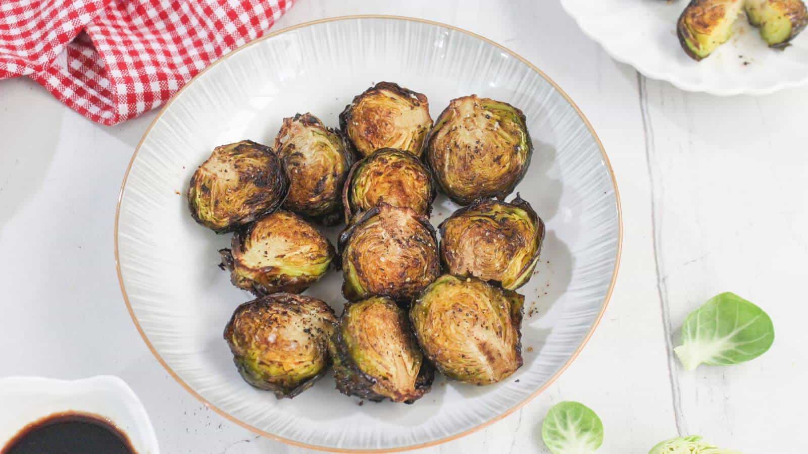 A white bowl filled with roasted Brussels sprouts, placed on a white surface with a red checkered cloth and scattered Brussels sprout leaves nearby.