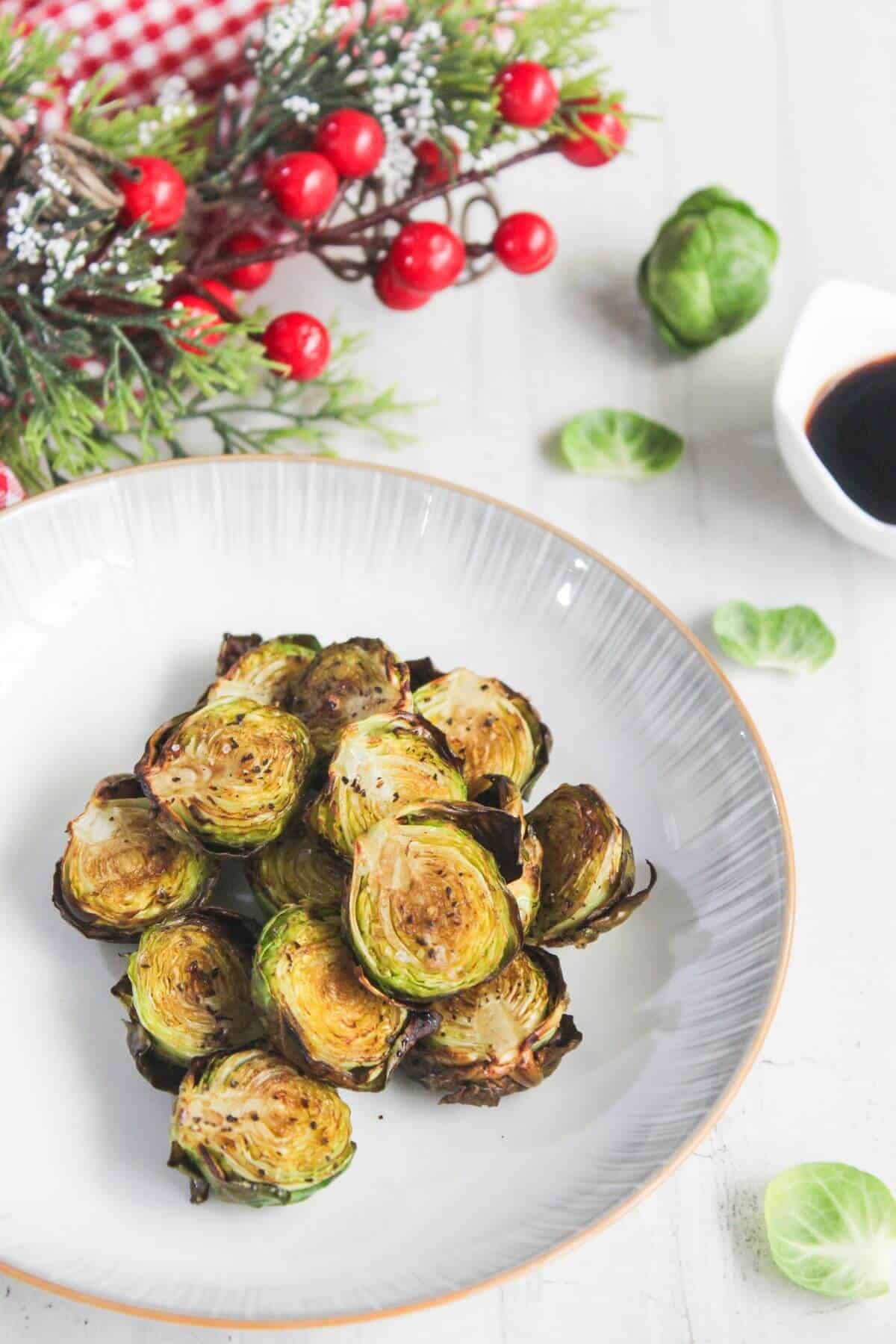 A plate of roasted Brussels sprouts on a white table, with a holiday decoration and a small bowl of dark sauce in the background.