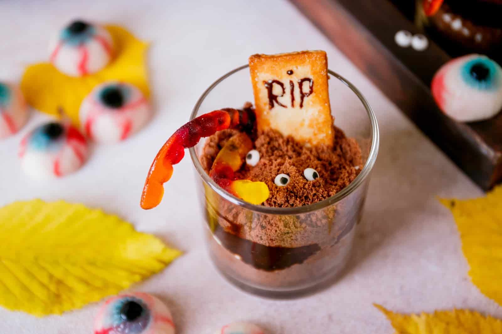 A Halloween-themed dessert in a glass, decorated with a cookie marked "RIP," candy eyes, chocolate crumbs, and a gummy worm, surrounded by fake eyeballs and yellow leaves.