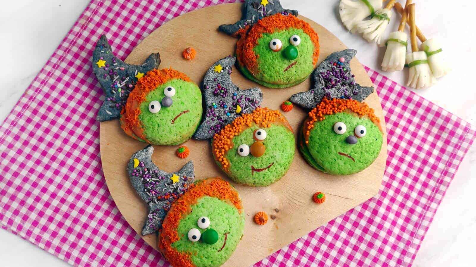 Six round cookies decorated as green-faced witches with orange hair and pointy hats on a wooden board, placed on a pink checkered cloth. Small broomstick-shaped treats are nearby.