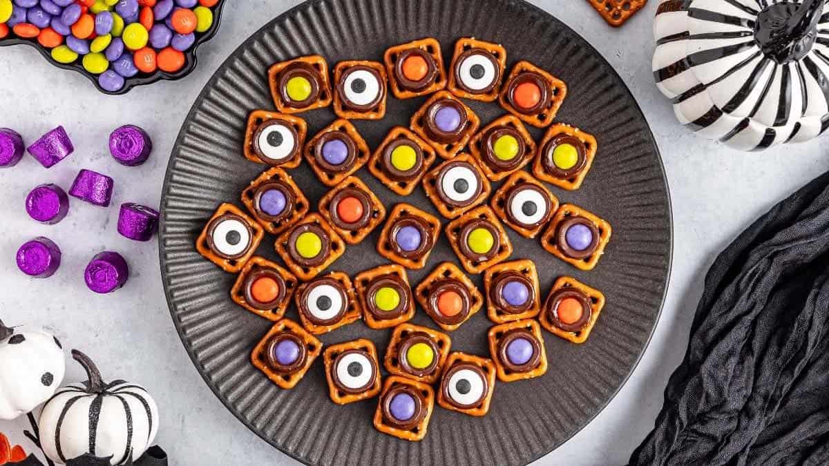 A black plate holds square pretzel snacks topped with colorful candies and candy eyes, surrounded by Halloween-themed decorations.