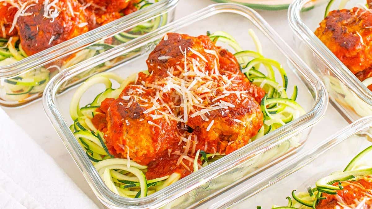 Glass containers filled with zucchini noodles, topped with meatballs in tomato sauce and grated cheese.