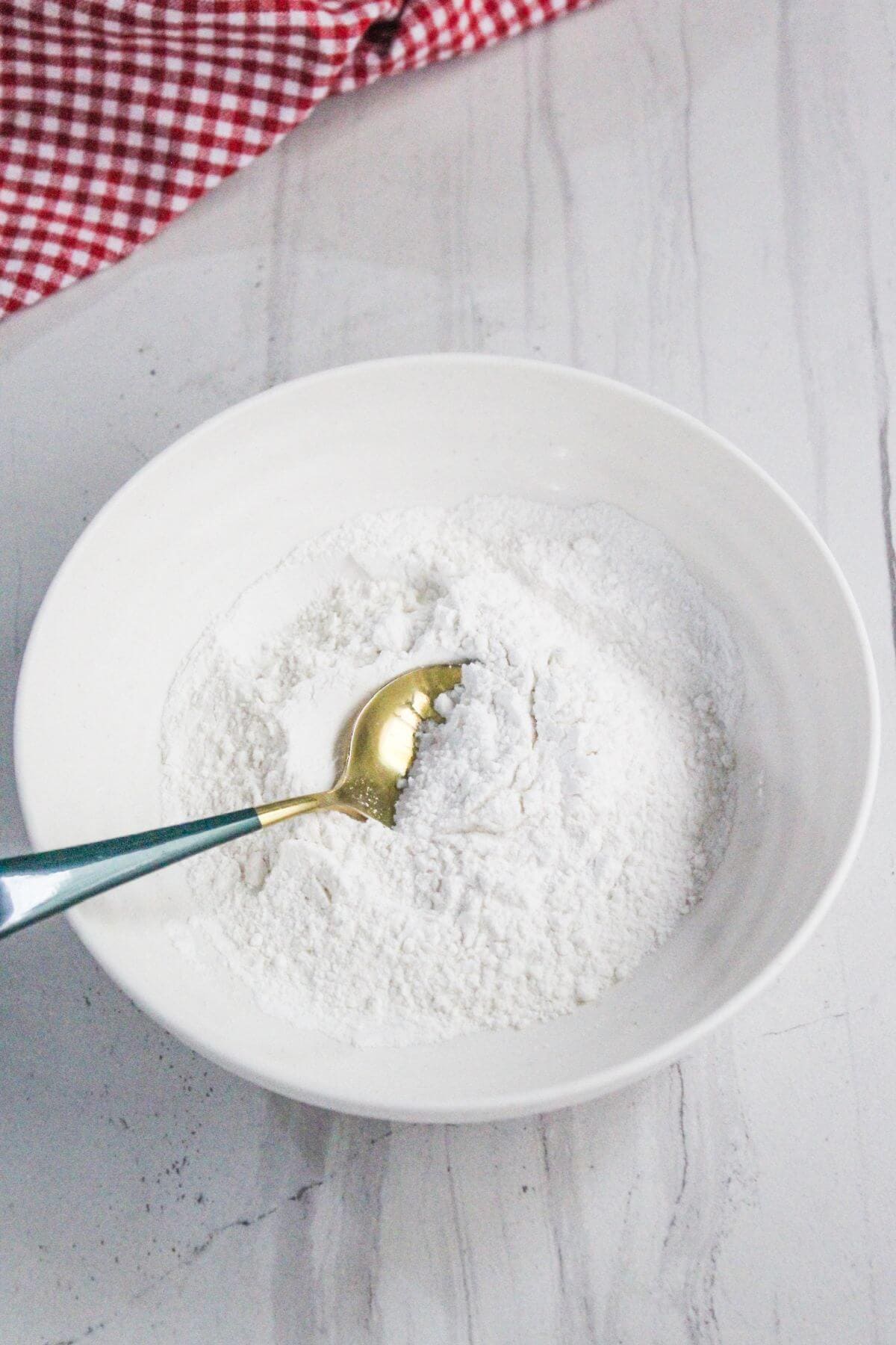 A white bowl filled with flour and a gold spoon on a white marble surface, with a red and white checkered cloth in the background.